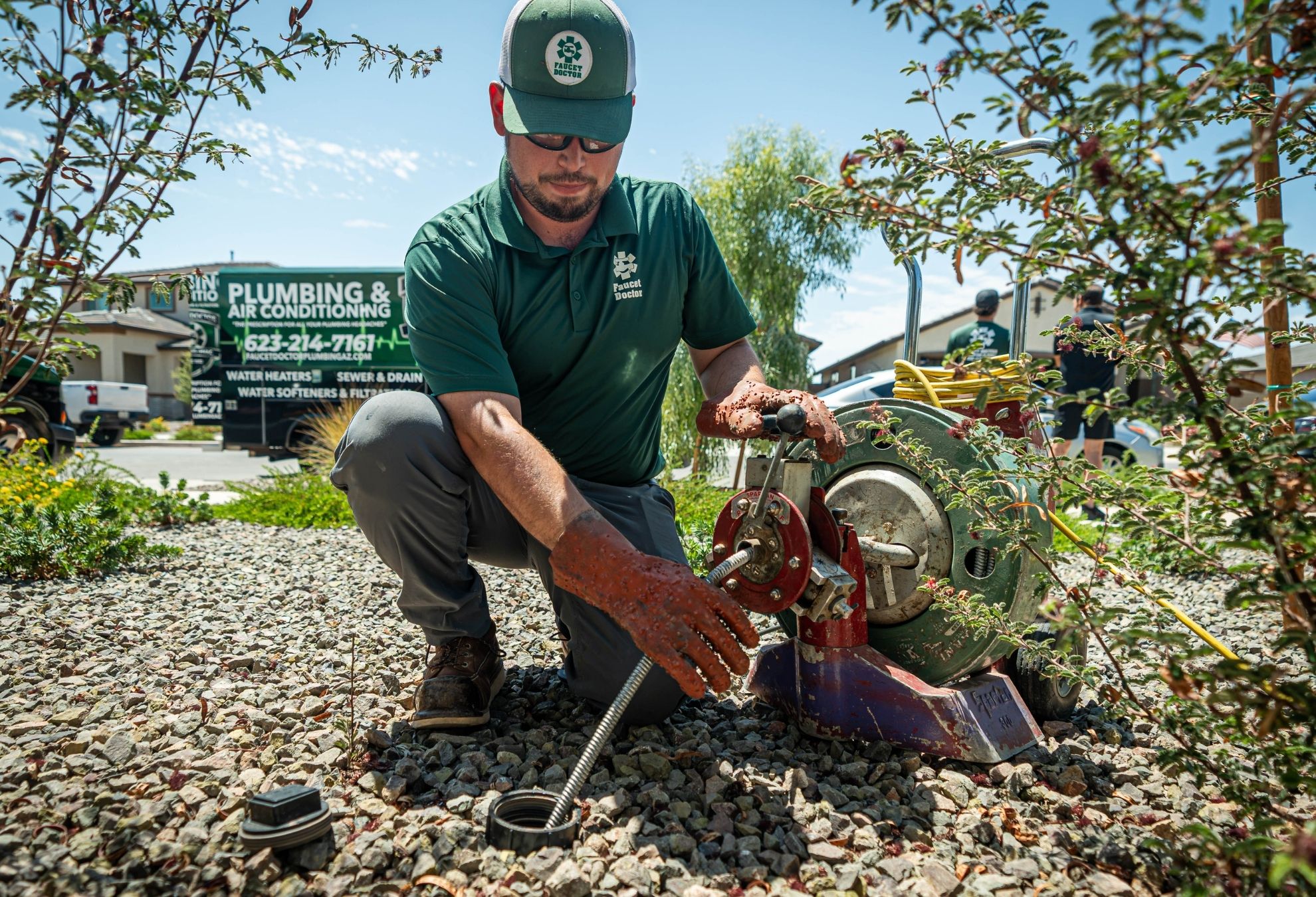 A plumber in a green shirt repairs a drain with a snake machine in a residential garden setting.